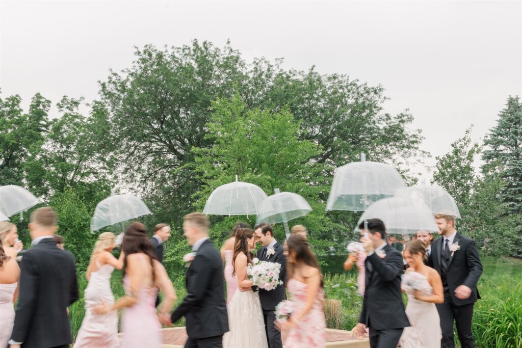 The bride and groom share a kiss while their bridal walks around them in a blur with clear umbrellas.