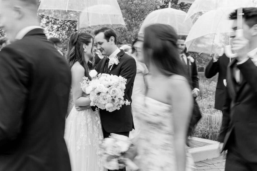 A black and white photo focused on the bride and groom while the bridal party walks around them with umbrellas.