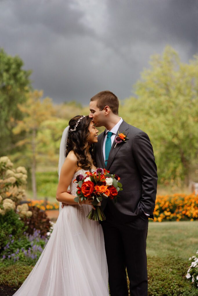 Tina and Connor kiss in the garden with colorful flowers and a stormy sky behind them.