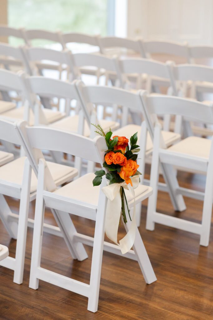 Orange and red flowers decorate a white ceremony chair along the indoor aisle.